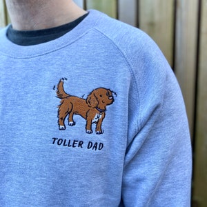 May include: A light blue sweatshirt with a brown dog embroidered on the front. The dog is looking to the right and has a collar with a tag. The text "TOLLER DAD" is embroidered below the dog.