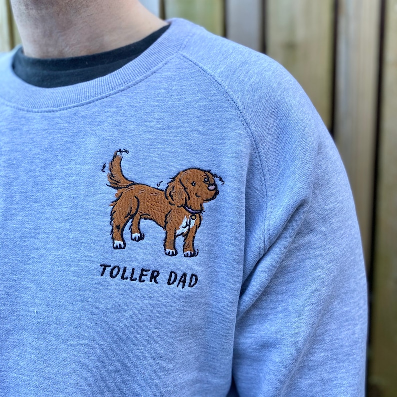 May include: A light blue sweatshirt with a brown dog embroidered on the front. The dog is looking to the right and has a collar with a tag. The text "TOLLER DAD" is embroidered below the dog.