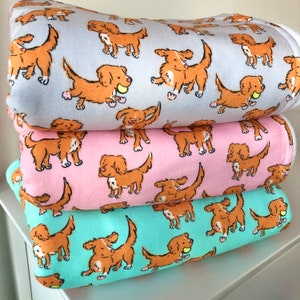 May include: Three soft blankets stacked, each with a repeating pattern of cartoon dogs in shades of brown and tan. The blankets are light blue, pink, and teal. The dogs are holding tennis balls.