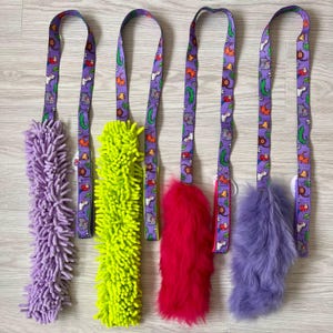 May include: Five dog toys with colorful patterned straps. The toys are a purple fuzzy rope, a green fuzzy rope, a pink fuzzy ball, a purple fuzzy tail, and a purple fuzzy tail.