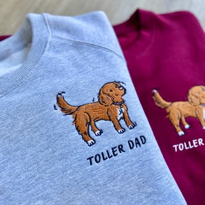 May include: Two sweatshirts, one gray and one burgundy, with embroidered images of a brown dog. The gray sweatshirt has the text "Toller Dad" embroidered below the dog. The burgundy sweatshirt has the text "Toller" embroidered below the dog.