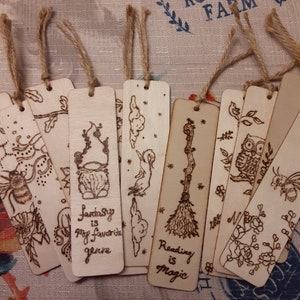 May include: A set of eight wooden bookmarks with various designs, including a spider, a cauldron, a cloud, a broom, an owl, and a flower. The bookmarks are tied with twine and have handwritten text, including "Fantasy is my favourite genre" and "Reading is magic".