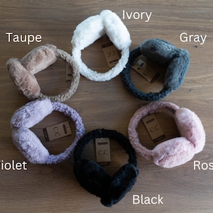 May include: Assortment of faux fur earmuffs in various colors, including ivory, taupe, gray, violet, rose, and black. Each earmuff has a brown tag with the letters "C.C."