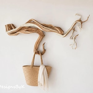 May include: A wall-mounted natural driftwood branch sculpture featuring a horizontal, twisted form with light and dark brown wood grain textures. A woven tan straw tote bag with a white fabric cloth draped over the handle hangs from the branch.