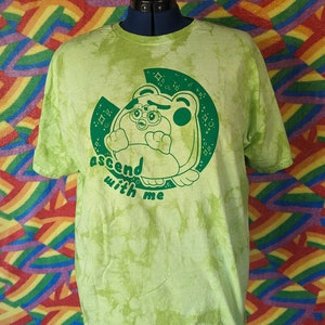 May include: Green tie-dye t-shirt with a green cartoon character wearing a crown and wings. The character is surrounded by stars and the text "ascend with me".