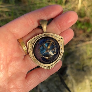 May include: A gold-toned pendant necklace with a shield-shaped frame. The pendant features a blue and teal orb with visible clockwork gears. The necklace is suspended from a black cord.