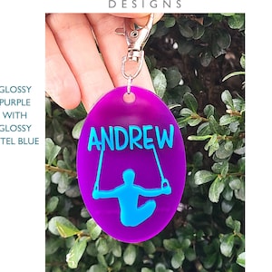 May include: A glossy purple keychain with a silver clasp. The keychain has the name "ANDREW" in blue letters, along with a blue gymnast figure. The text "SAKU DESIGNS" is at the top.
