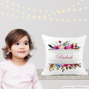 Puede incluir: Una almohada blanca con un diseño floral y texto en hebreo. El texto dice: "Que el que bendijo a nuestros antepasados bendiga a esta niña, y que su nombre en Israel sea llamado Rivkah. Así como ella ha entrado en el pacto, que ella entre en una vida de Torá, relaciones amorosas y bondad."