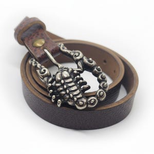 May include: A brown leather belt with a silver scorpion buckle. The belt is rolled up, showcasing the buckle's intricate details. The buckle is a detailed scorpion design, adding a unique touch to the accessory.