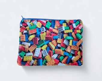 May include: Rectangular zippered pouch featuring a colorful pattern of toy building blocks. The design includes red, yellow, green, blue, and white blocks. The pouch has a blue zipper and a blue border.