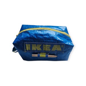 Könnte beinhalten: Eine blaue Reißverschlusstasche mit gelbem Reißverschluss und dem Wort "IKEA" in gelben Blockbuchstaben. Die Tasche besteht aus einem gewebten Material und hat einen kleinen Holzakzent.