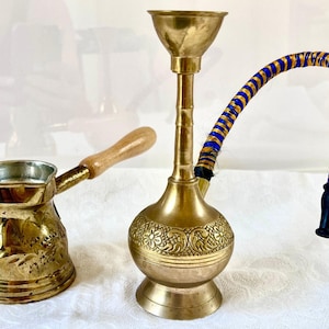 May include: A brass coffee pot with a wooden handle and a brass hookah with a blue and gold striped hose. The coffee pot features etched floral patterns on its body, while the hookah has a tall neck and a wide bowl with engraved details.
