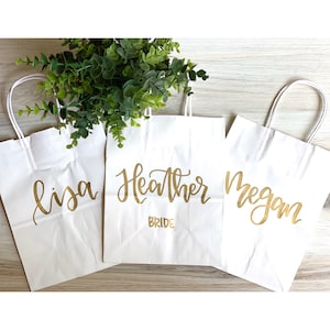 May include: Three white paper gift bags with gold lettering. The bags are labeled "Lisa", "Heather Bride", and "Megan".