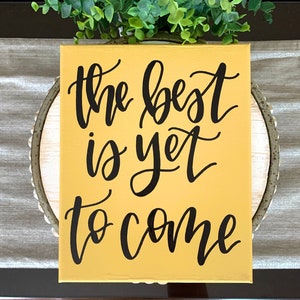 May include: A yellow canvas sign with black calligraphy that reads "The best is yet to come."