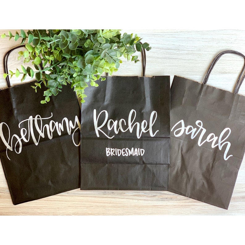 May include: Three black paper gift bags with white lettering. The bags are labeled "Bethany", "Rachel Bridesmaid", and "Sarah".