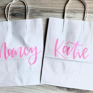 May include: Two white paper gift bags with pink lettering. The bag on the left says "Nancy" and the bag on the right says "Katie".