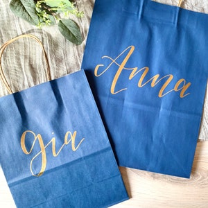 May include: Two blue paper gift bags with gold lettering. The larger bag says "Anna" and the smaller bag says "Gia".