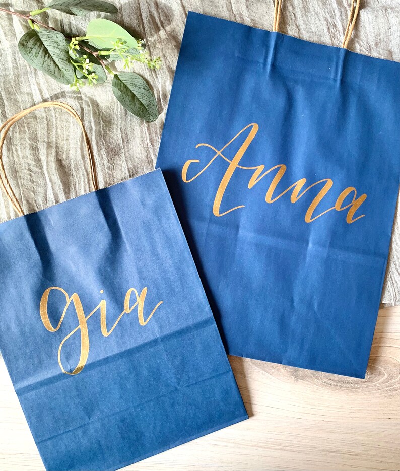 May include: Two blue paper gift bags with gold lettering. The larger bag says "Anna" and the smaller bag says "Gia".