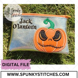 May include: A silver zippered pouch with a black and green embroidered Jack O'Lantern with a stitched-on grin. The text "Jack O'Lantern" is embroidered in black above the pumpkin.