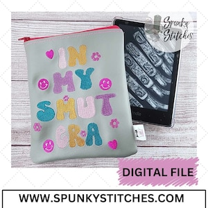 May include: A gray zippered pouch with the text "IN MY SHUT ERA" embroidered in colorful letters and smiley faces. The pouch is sitting on a wooden surface next to a black and white e-reader.