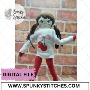 May include: A white doll sweater with a pink bow and embroidered cherries. The sweater is on a doll with brown hair and red pants. The doll is standing in front of a brick wall.