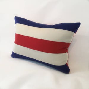 May include: A rectangular pillow with a striped pattern. The pillow features alternating bands of red, white, and blue. The pillow's edges are a solid blue color. The pillow is a decorative item.