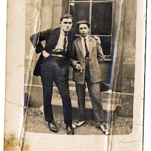 Peut inclure: Deux hommes en costume se tiennent devant un bâtiment. L'homme de gauche a la main dans sa poche et regarde l'appareil photo. L'homme de droite tient une cigarette et regarde sur le côté. Le bâtiment a une fenêtre avec une enseigne qui dit "The E Res".