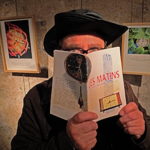 Op de afbeelding: Een persoon met een zwarte hoed en bril houdt een boek vast met de titel "Les Matins" van Sylvain Soulard. De omslag van het boek toont een klok en een gebouw.