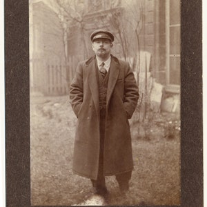 Può includere: Un uomo con un cappotto scuro e un berretto sta in piedi davanti a un edificio. Sta guardando la telecamera. La foto è in bianco e nero.