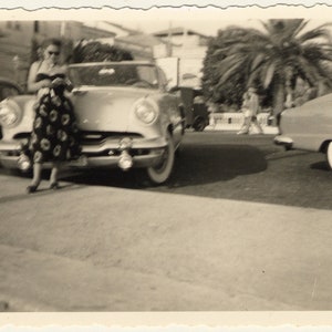 Peut inclure: Une photo en noir et blanc d'une femme debout devant une voiture ancienne. La voiture est une berline de couleur claire avec des accents chromés. La femme porte une robe à fleurs et regarde la caméra.