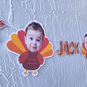 May include: A festive banner featuring a child's face incorporated into a turkey design. The banner includes the name "JACK" in patterned letters. The turkey's feathers are in shades of orange and yellow, with a brown body.