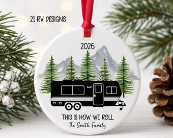 May include: A round white ceramic ornament featuring a black recreational vehicle graphic, green pine trees, and blue mountains. The text reads 2026, THIS IS HOW WE ROLL, and The Smith Family. A red ribbon is attached to the top for hanging.