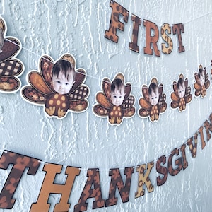 May include: Thanksgiving banner with the words "FIRST THANKSGIVING" and baby turkey decorations. The letters are brown with a floral pattern and leopard print accents. The background is a textured light blue wall.
