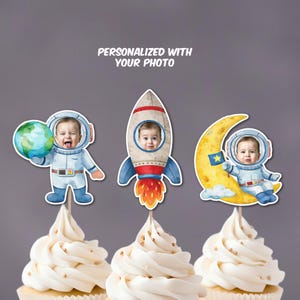 May include: Three cupcake toppers featuring a baby's face in astronaut-themed designs. One holds a globe, another is in a rocket, and the third is on a crescent moon. The toppers are on cupcakes with white frosting and gold sprinkles. Text at the top reads "PERSONALIZED WITH YOUR PHOTO."