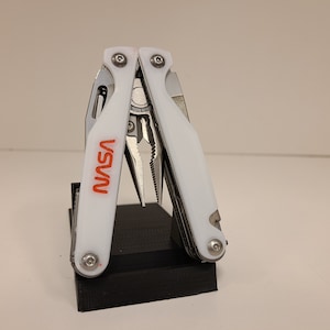 May include: A white multi-tool with a red "NASA" logo. The tool is open and has pliers, a knife, and other tools.