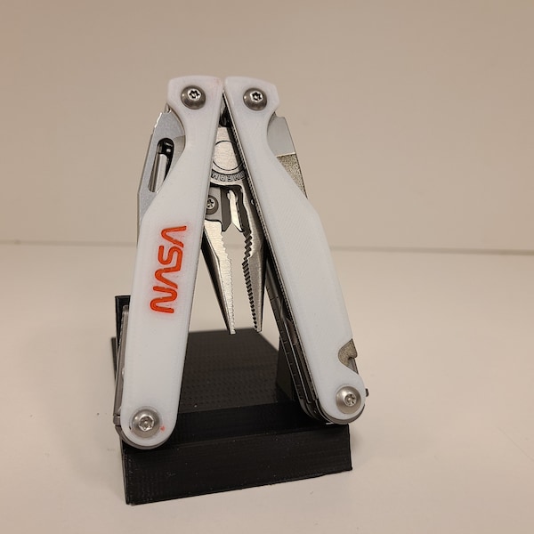 May include: A white multi-tool with a red "NASA" logo. The tool is open and has pliers, a knife, and other tools.