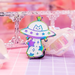 May include: A rainbow iridescent enamel pin of a cat in a UFO with stars and glitter. The pin is white with a black outline.