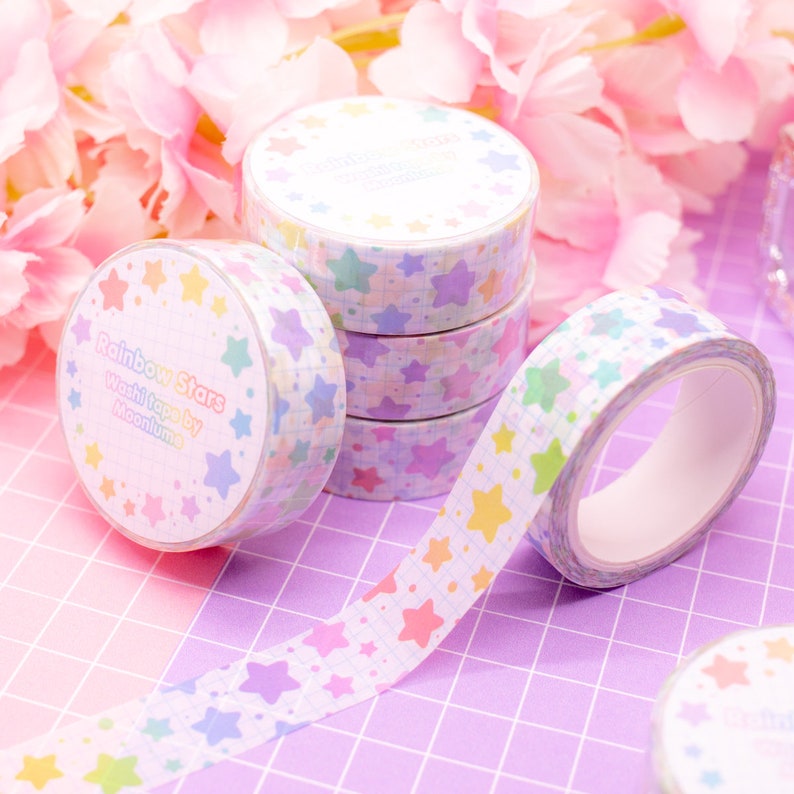 May include: A set of four rolls of rainbow star patterned washi tape. The tape is white with a grid pattern and colorful stars. The text on the tape reads "Rainbow Stars Washi Tape by MoonLume".