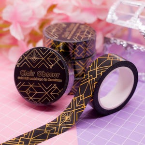 Könnte beinhalten: Schwarzes Washi-Tape mit einem goldenen geometrischen Muster. Auf dem Tape steht "Clair Obscur" und "Gold foil washi tape by Moonlume". Mehrere Rollen sind gestapelt, eine davon abgerollt, das goldene Muster zeigend.