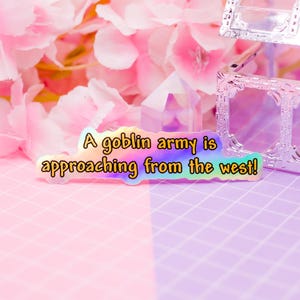 Könnte beinhalten: Ein holografischer Aufkleber mit dem Text "A goblin army is approaching from the west!" in einer verspielten Schriftart. Der Aufkleber hat einen Regenbogen-Schimmer und einen gelben Rand, vor einem rosa-lila Hintergrund.