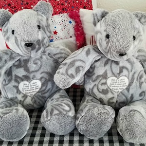 May include: Two gray plush teddy bears with a swirled pattern. Each bear has a heart-shaped patch with text. The bears are on a black and white checkered surface, with decorative pillows in the background.