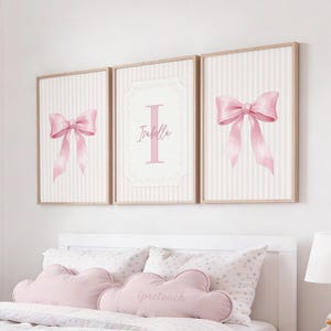 Pode incluir: Três quadros emoldurados com desenhos de laços rosa sobre um fundo listrado. A moldura central apresenta a letra "I" e o nome "Isabella". As molduras têm um acabamento em madeira clara, criando uma estética suave e feminina.