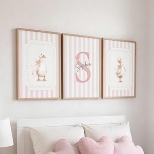 Può includere: Tre stampe incorniciate con finitura in legno chiaro. Due stampe presentano un'oca ad acquerello con un fiocco rosa, su uno sfondo a righe rosa e bianche. La stampa centrale mostra la lettera "S" e il nome "Sophia" in rosa.