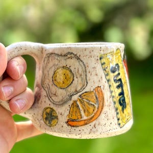 May include: A white ceramic mug with a speckled glaze. The mug has a handle and is decorated with hand-painted illustrations of a fried egg, a slice of orange, and a stick of butter.