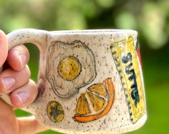 May include: A white ceramic mug with a speckled glaze. The mug has a handle and is decorated with hand-painted illustrations of a fried egg, a slice of orange, and a stick of butter.