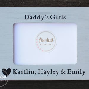 以下が含まれることがあります： 「Daddy's Girls」と「Kaitlin, Hayley & Emily」の文字が印刷された薄い青色の木製写真フレーム。フレームの左側にハート型の切り抜きがあり、中央には「Heartfelt by Heather」の小さなロゴがあります。