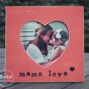 May include: A pink wooden picture frame with a heart-shaped cutout. The frame has the words "mama love" printed on the bottom. A photo of a mother and daughter is visible through the cutout.