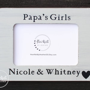 Könnte beinhalten: Ein rechteckiger, weißer Bilderrahmen mit Used-Look, mit den Worten "Papa's Girls" oben und "Nicole & Whitney" unten. Ein schwarzes Herz befindet sich neben "Whitney". Der Rahmen hat einen weißen Innenraum und ein Logo in der Mitte.