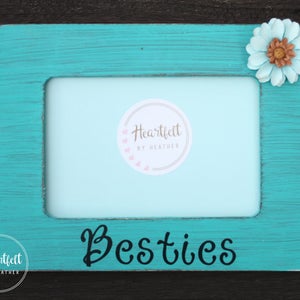 May include: Teal blue wooden picture frame with a white flower and the word "Besties" in black lettering. The frame has a white mat and a clear window for a photo.