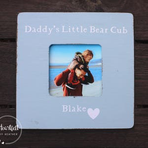 May include: A light blue wooden picture frame with a white heart and the text "Daddy's Little Bear Cub" and "Blake". The frame has a photo of a father and son, with the son on the father's shoulders.